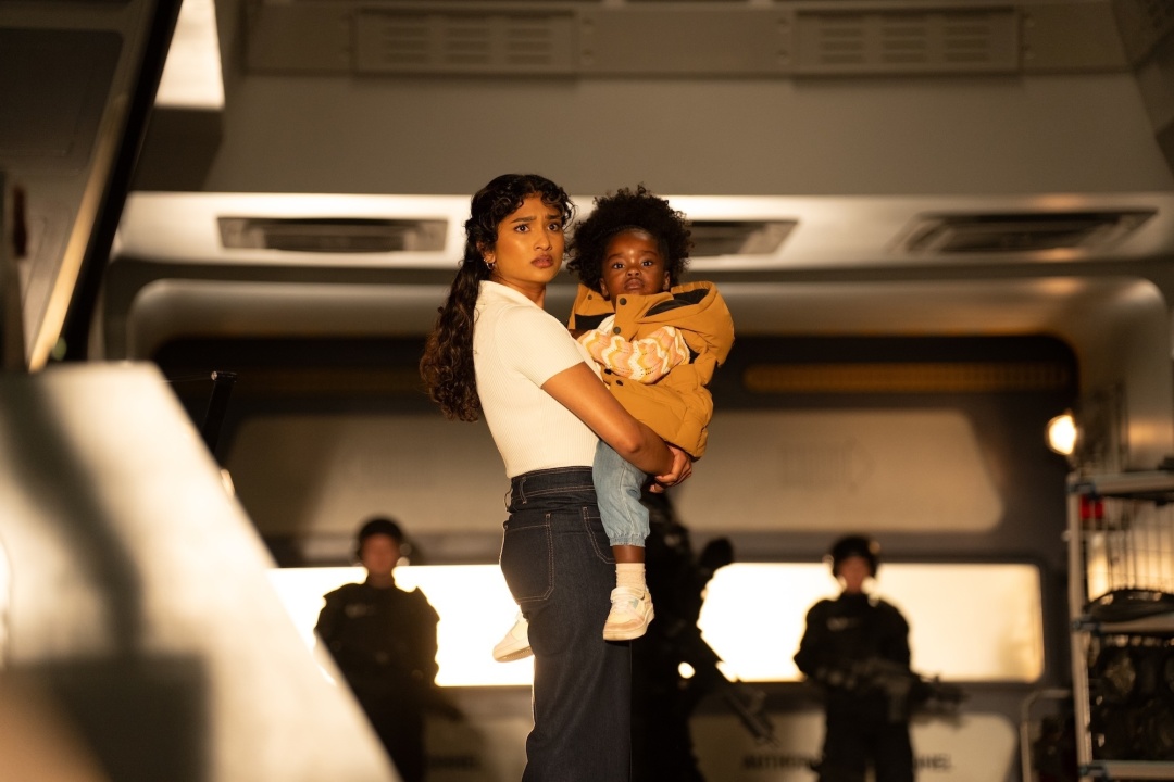 A woman in a white shirt and jeans holds a young child in a yellow jacket, standing in a room with armed guards in the background.