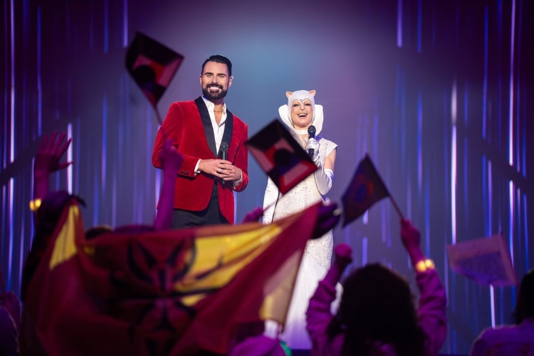 A man in a red blazer stands next to a person in a white animal costume on stage, while an audience waves flags in the foreground.