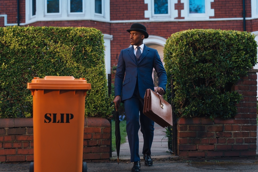 A man in a suit and bowler hat carries a briefcase while exiting a gate near an orange bin labeled "SLIP," in front of a brick building with hedges.
