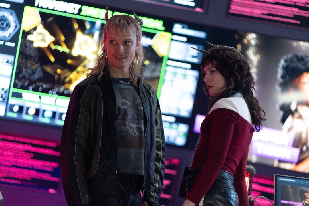 Two people in futuristic attire stand in a control room with colorful screens displaying data and graphics in the background.