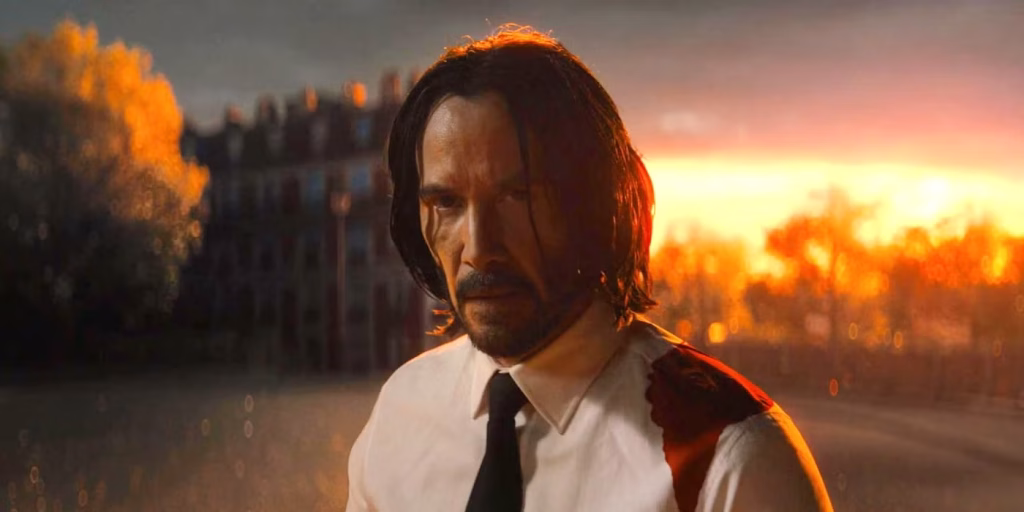 A man with long hair and a beard, wearing a white shirt and black tie stained with blood, stands outdoors at sunset with trees and a building in the background.