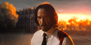A man with long hair and a beard, wearing a white shirt and black tie stained with blood, stands outdoors at sunset with trees and a building in the background.