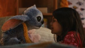 A young girl in a red shirt sits closely facing a blue, furry alien creature with large ears and black eyes in a cozy indoor setting.