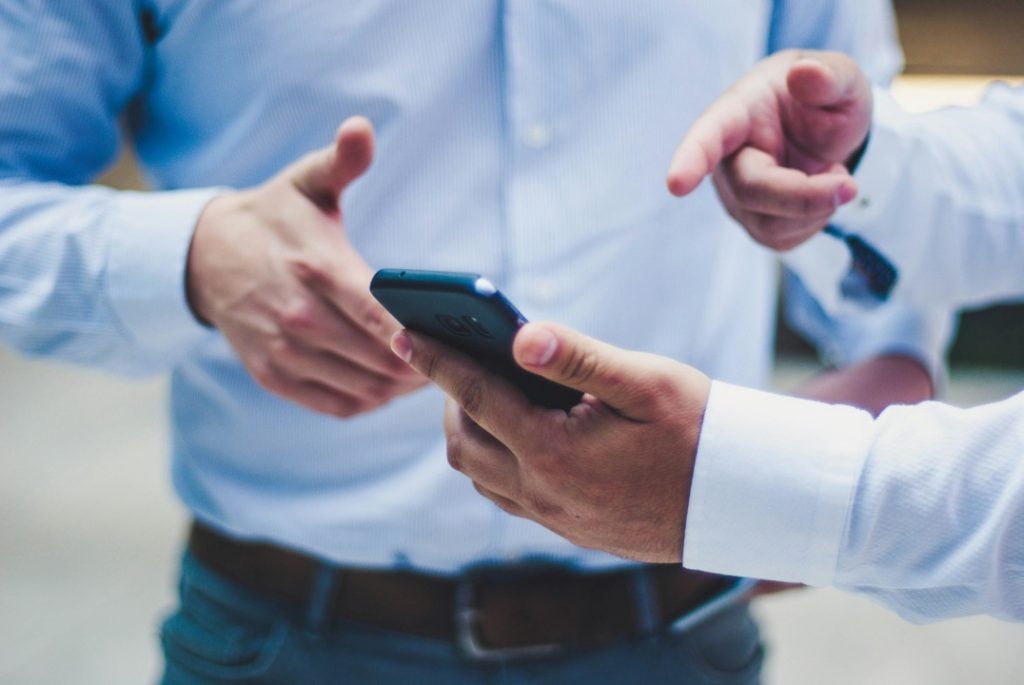 Two people in button-up shirts discuss something on a smartphone, with one person holding the phone and the other gesturing and pointing at the screen.