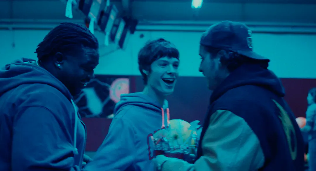 Three people in casual clothing stand together indoors, two facing a third person who is smiling, with decorations visible in the background.