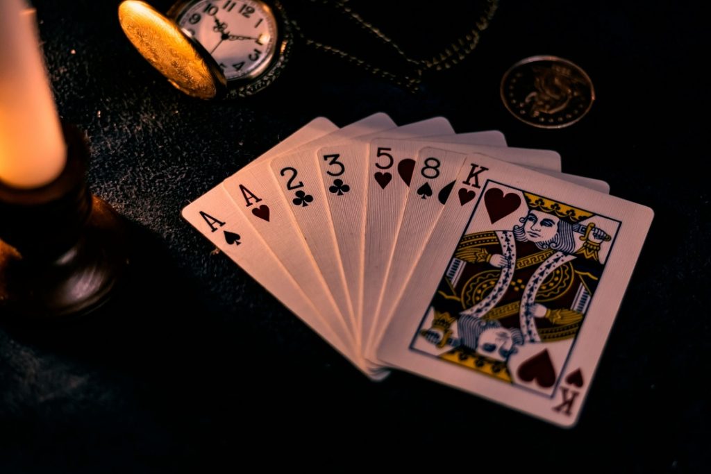 A fan of six playing cards—Ace, 2, 3, 5, 8 of clubs, and King of hearts—lies on a dark surface next to a pocket watch and a coin.