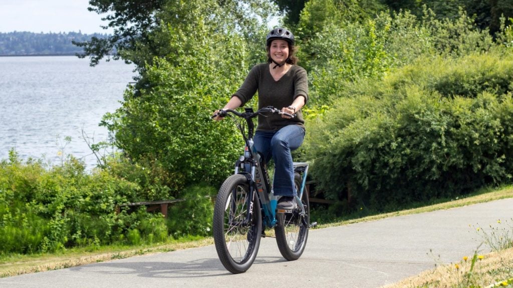 A person wearing a helmet rides a bicycle on a paved path beside a body of water, surrounded by green trees and bushes.