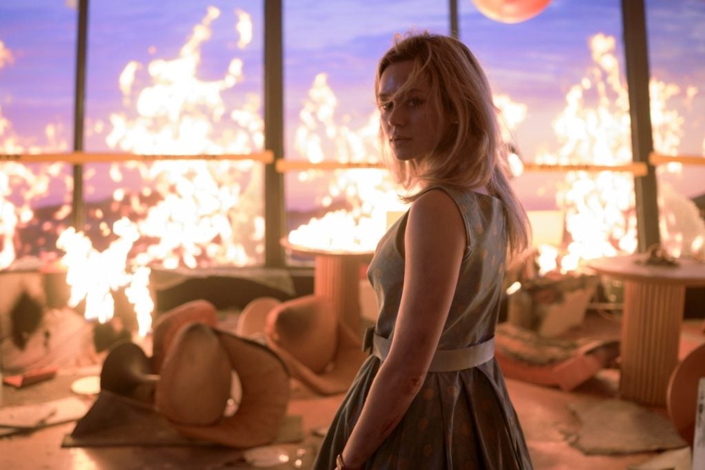 A woman in a dress stands in a room filled with overturned furniture and intense flames, with large windows in the background.