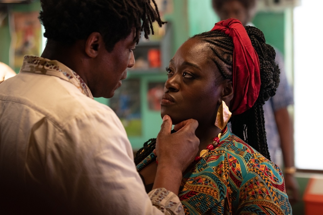 A man gently holds a woman's chin while they look at each other closely in a colorful, indoor setting.