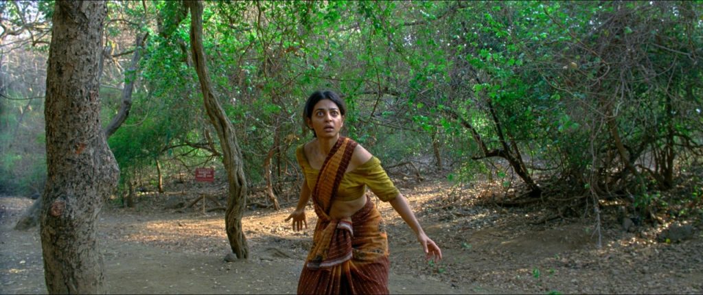 A woman in a traditional sari stands alert and cautious in a forested area, looking toward something off-camera.