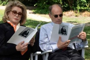 Two people sitting outdoors on white chairs, both reading magazines with the same black-and-white cover photo of a woman in formal attire. Greenery is visible in the background.