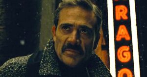 A man with a mustache stands outside at night, wearing a coat, with a neon sign reading "DRAGO" illuminated in the background.