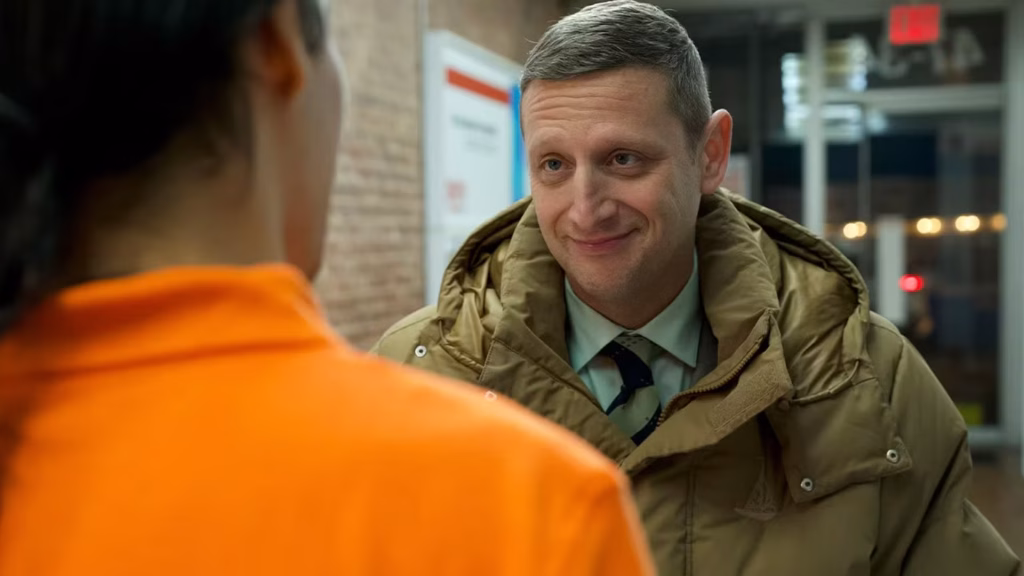 A man in a tan winter coat and tie smiles while talking to a person in an orange shirt indoors near a brick wall.