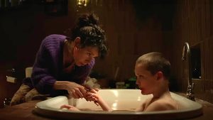 A24 To Release The Disturbing Horror Film ‘Bring Her Back’ On 4K UHD Blu-Ray This August A woman in a purple sweater tends to a young boy sitting in a bathtub, holding his hand in a dimly lit bathroom.