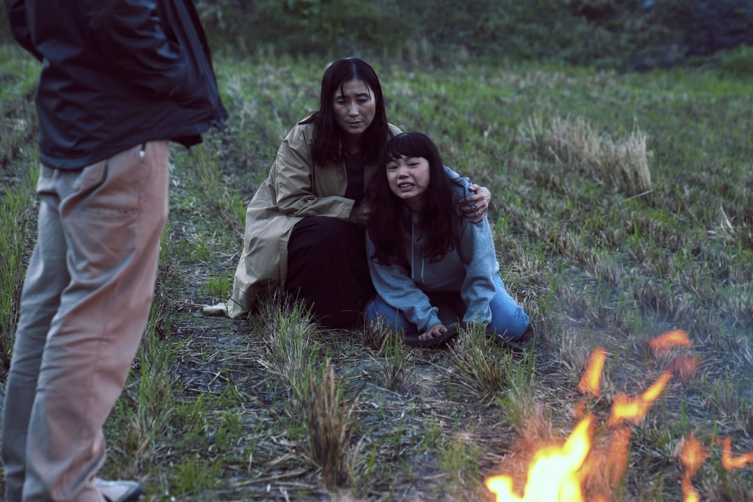 Two women sit on grass near a small fire; one is kneeling and appears upset while the other comforts her. A third person stands nearby, partially out of frame.
