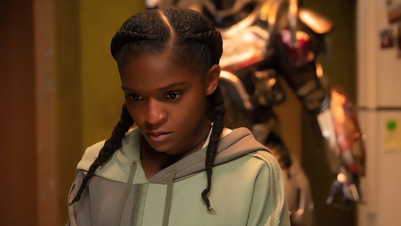 A young woman with two braids, wearing a light green hoodie, stands indoors with a serious expression. A metallic suit is visible in the background.