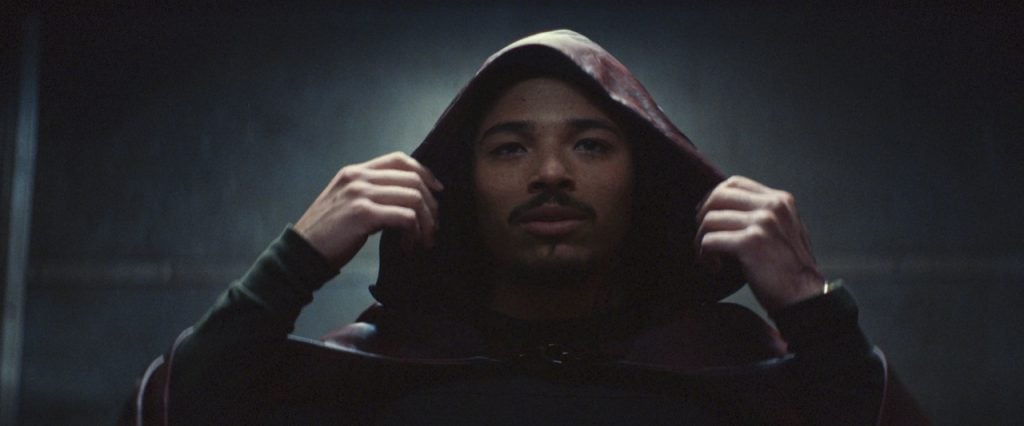 A person with a mustache and goatee pulls up the hood of a dark cloak, looking forward in a dimly lit space.