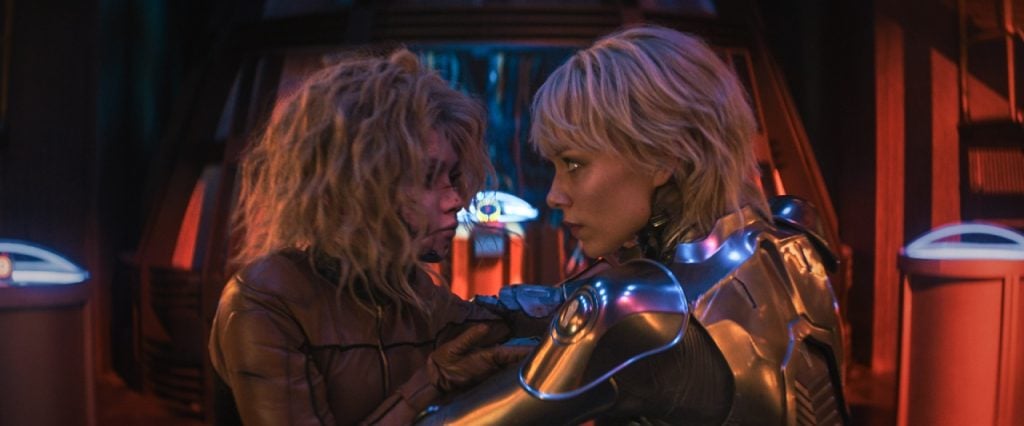 Two women in futuristic suits face each other closely in a dimly lit, high-tech environment with vibrant red and blue lighting.