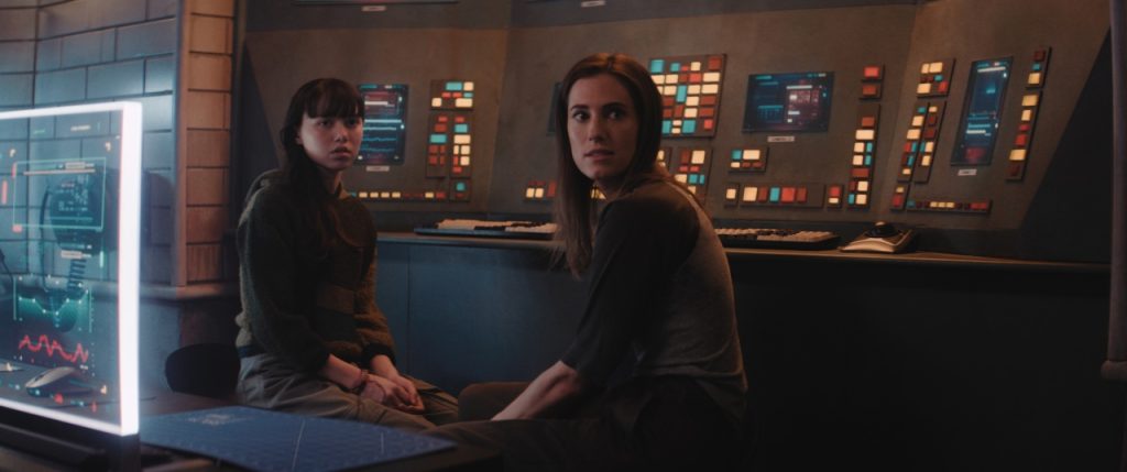 Two women sit in front of a large control panel with numerous buttons and screens, looking back over their shoulders with concerned expressions.