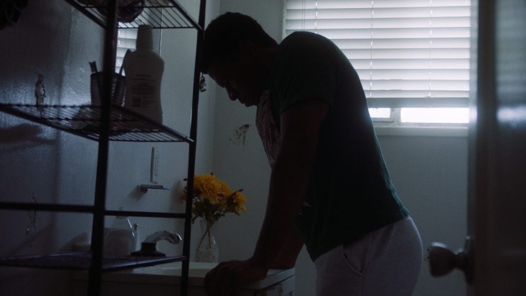 A person leans over a bathroom sink with their head down, next to a vase of flowers, under a window with closed blinds in dim lighting.
