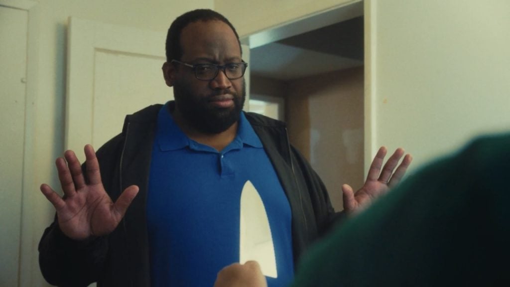 A man in a blue shirt stands with his hands raised while another person points a large knife at him in a room.