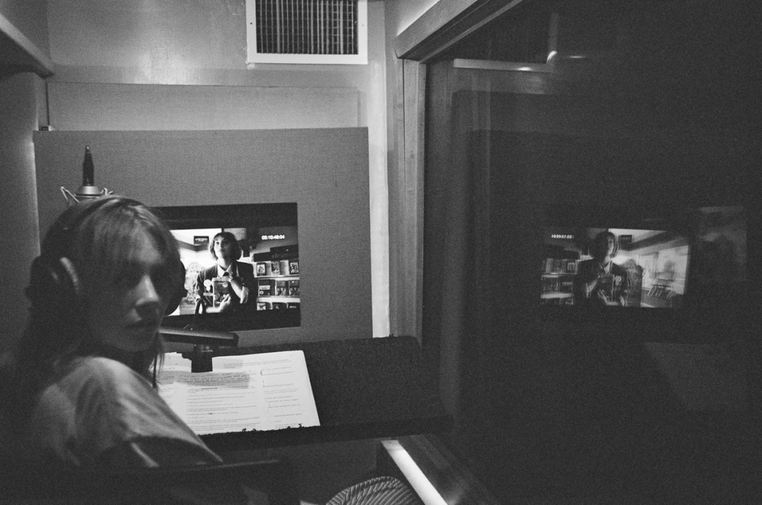 A person wearing headphones sits in a recording booth with a microphone, looking back at the camera; a script and a screen displaying a film scene are in front of them.