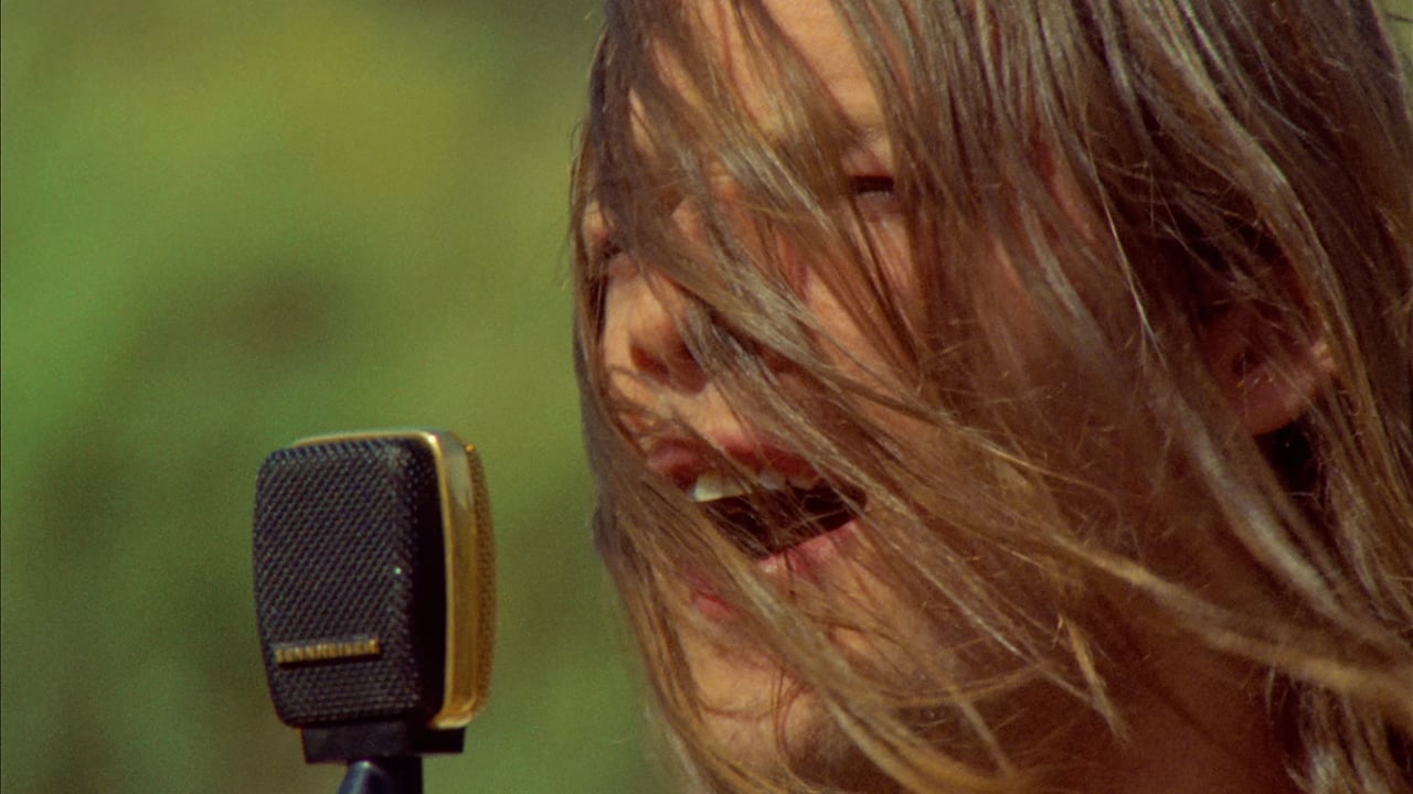 A person sings into a vintage microphone with hair partially covering their face, set against a blurred outdoor background.