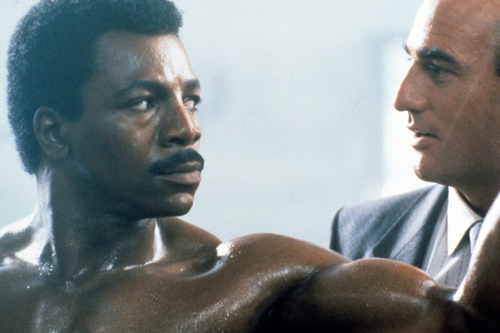 A muscular man with a mustache looks intently at a man in a suit who is speaking to him in close proximity.