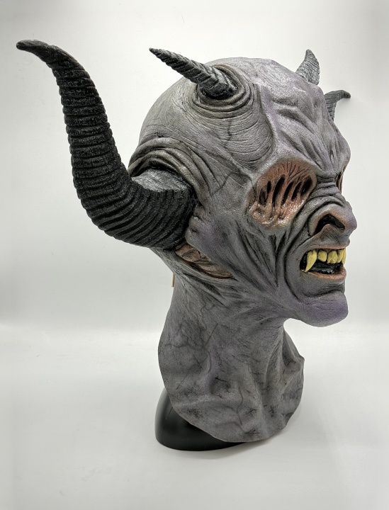 A detailed latex mask of a demonic creature with gray skin, large twisted black horns, pointed ears, yellow fangs, and deep facial wrinkles, shown on a plain white background.