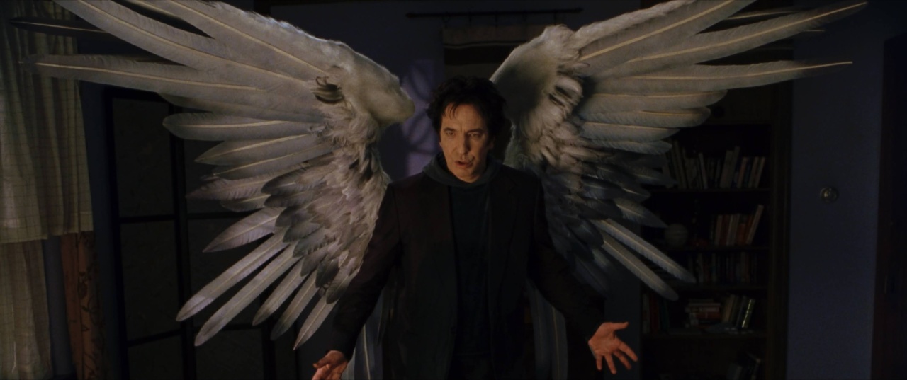A man stands indoors with large, outstretched, feathered wings attached to his back, wearing dark clothing and looking forward with an intense expression.