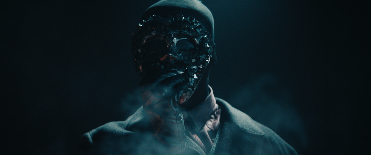 A person wearing a reflective, gem-encrusted mask and dark clothing stands in mist or smoke, holding their hand near their face.