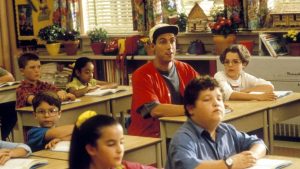 A man wearing a red shirt sits at a classroom desk among young children, all facing forward and appearing to listen or read.