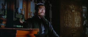 Eureka Entertainment Announces Summer Blu-Ray Titles From Lo Wei, Jimmy Wang Yu & More A man with a long beard and traditional attire sits on an ornate wooden chair in a dimly lit room with decorative wooden panels.