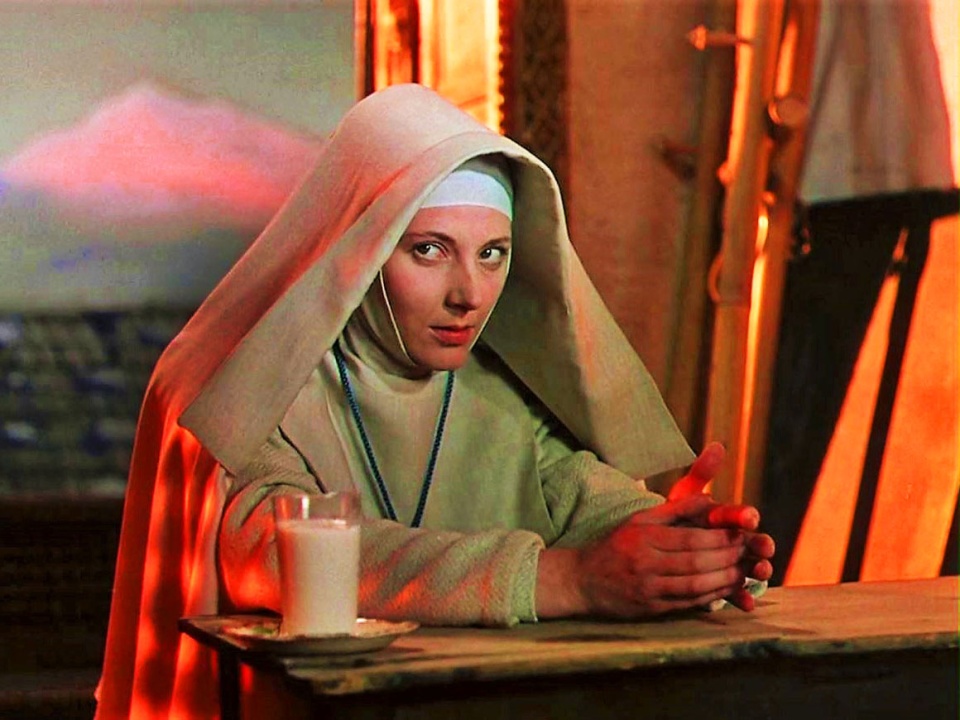 A woman dressed as a nun sits at a table with a glass of milk, hands clasped, in a warmly lit room with an orange glow.