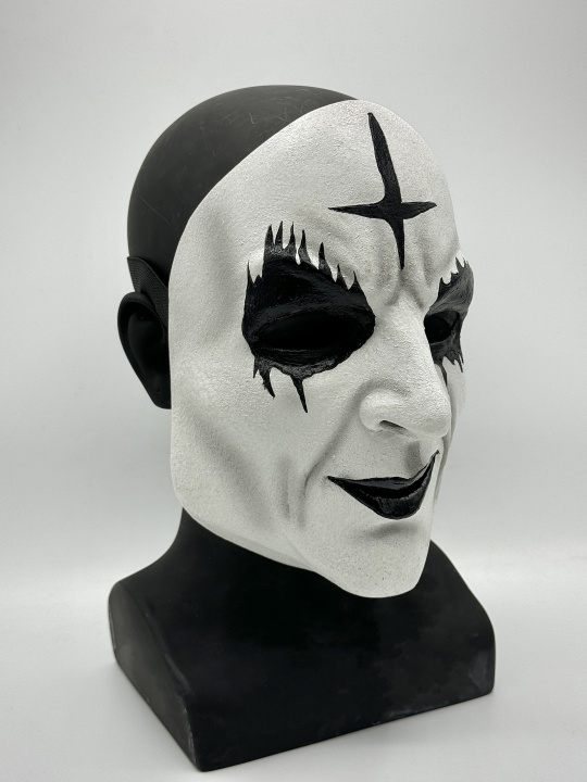A black and white mask with a painted cross on the forehead and dramatic black eye and lip makeup, displayed on a black bust against a plain background.