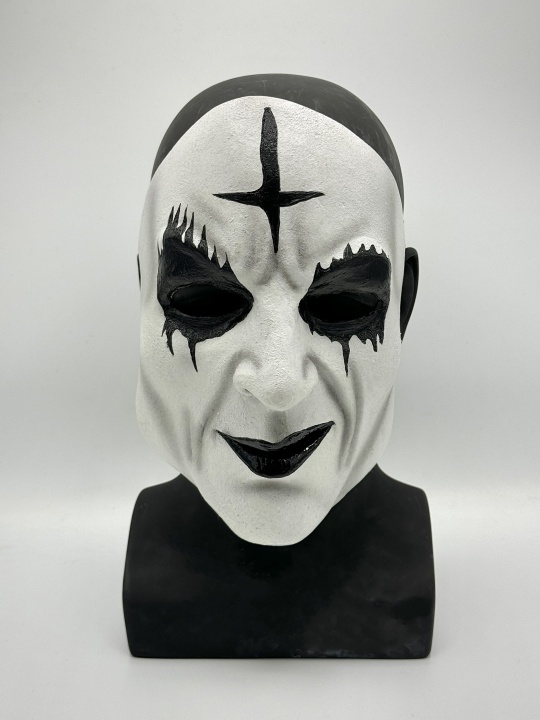 Bust of a head wearing a white mask with black eye and lip makeup, and a large black cross painted on the forehead, set against a plain background.