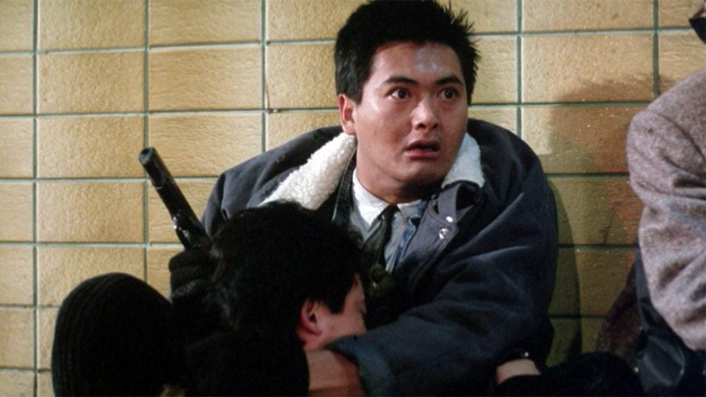 A man in a jacket holds a pistol while sitting against a tiled wall, appearing alert and tense, with another person leaning against him.
