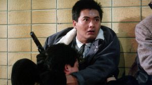 Shout! & Hong Kong Cinema Classics To Bring Chow Yun-Fat Favorite ‘City On Fire’ To 4K UHD Blu-Ray This August A man in a jacket holds a pistol while sitting against a tiled wall, appearing alert and tense, with another person leaning against him.