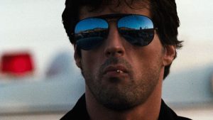 A man wearing reflective sunglasses and a toothpick in his mouth stares ahead, with a blurred background and reflections visible in his lenses.