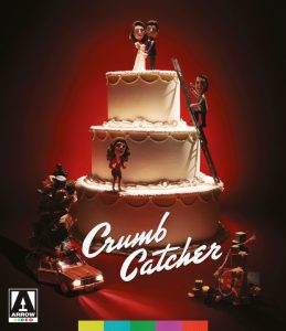 A two-tier wedding cake with three figurines on top; one man stands on a ladder, a woman stands on the cake, and another woman stands beside a car with boxes. "Crumb Catcher" is written in white.
