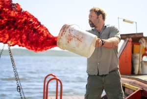 A man on a boat pours a large bucket of red liquid into the water, with the liquid forming a dramatic splash.