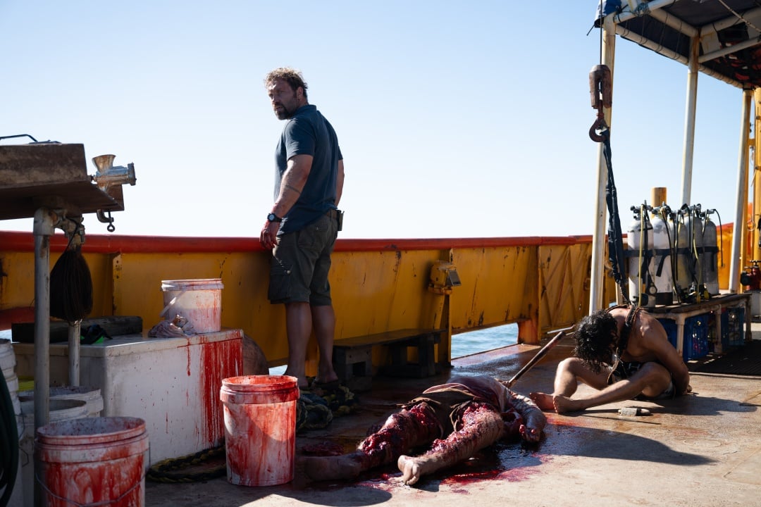 A man stands on a bloodstained boat deck beside a body, while another person sits on the floor nearby; diving equipment and storage containers are visible.