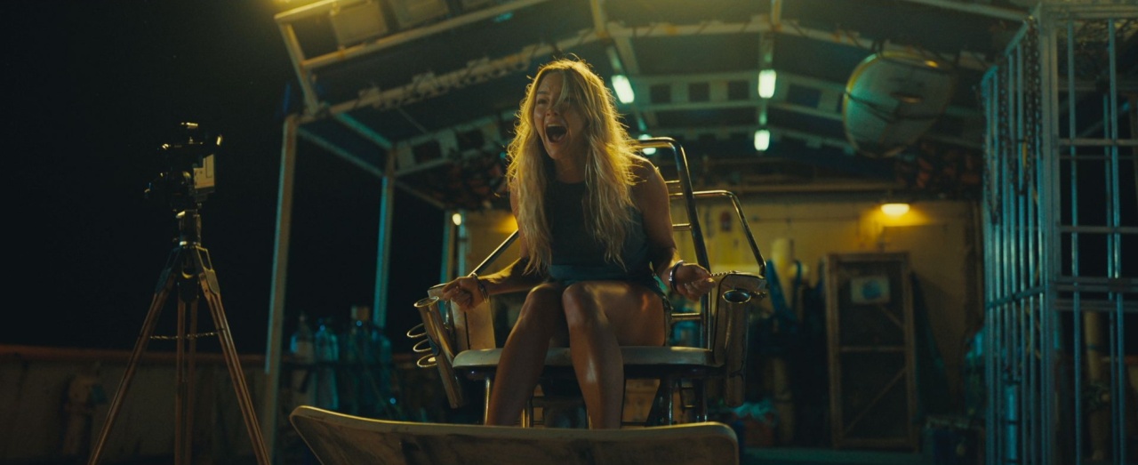 A woman with long hair sits restrained in a metal chair, screaming, inside a dimly lit industrial setting with equipment and a camera on a tripod nearby.