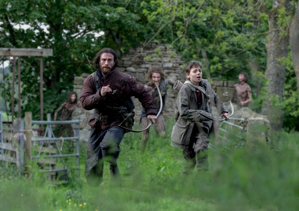 Two people run through grass in outdoor medieval-style clothing, chased by several others in the background, with trees and an old stone structure behind them.