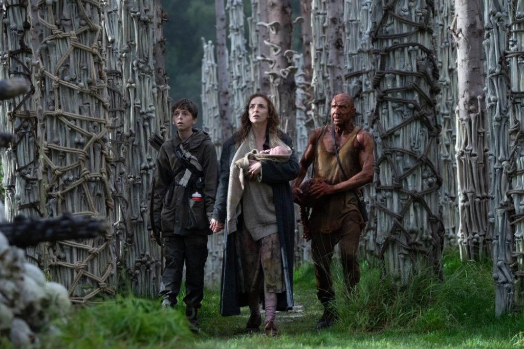 Three people, one holding a baby, walk through a forest of tall trees with intricate rope patterns tied around the trunks.