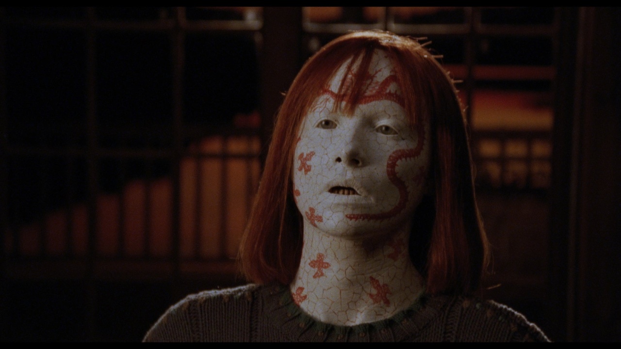 A person with pale, cracked skin covered in red, flower-like patterns and straight red hair stares ahead with a blank expression.