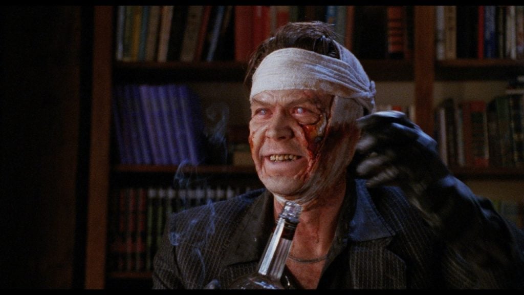 A man with a bloody, bandaged head and facial injuries smiles while holding a bottle and wearing a black glove; bookshelves are in the background.