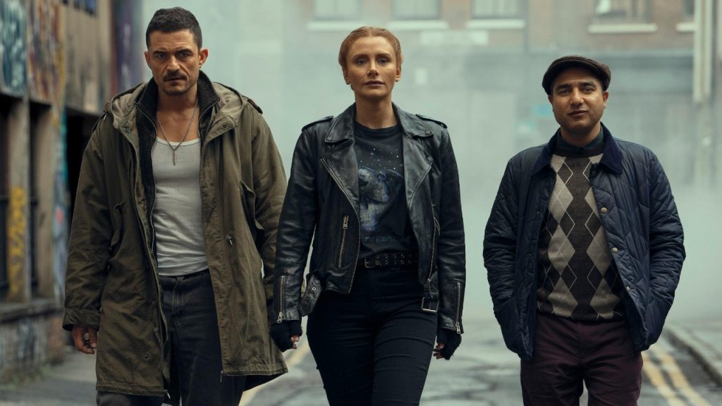 Three people walk side by side down a foggy urban street, wearing casual and rugged clothing, with serious expressions.