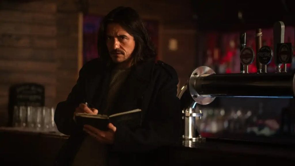 A man with long dark hair and a mustache stands at a bar, wearing a dark coat and writing in a notebook under dim lighting.