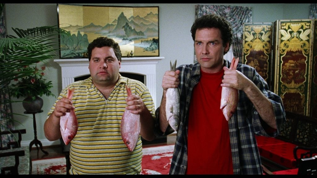 Two men stand indoors, each holding two whole fish by the tails. The man on the left wears a yellow striped shirt; the man on the right wears a red shirt and plaid overshirt.
